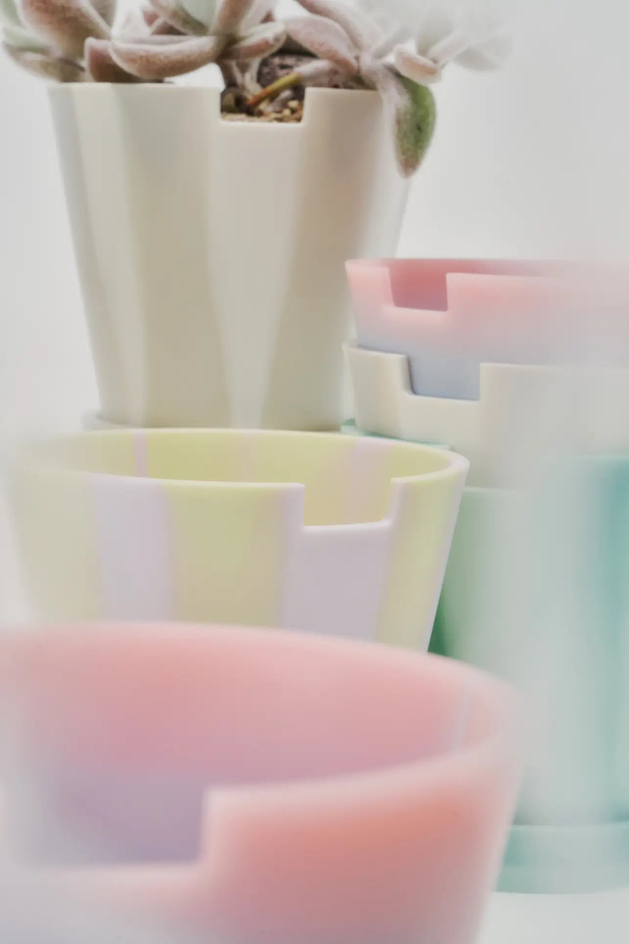 A soft-focus image displays several Plastic Ark Dirty Pots by Plastic Ark in pastel colors, some stacked with geometric edges. A plant with fuzzy leaves sits in the top pot, creating a light and airy vibe.