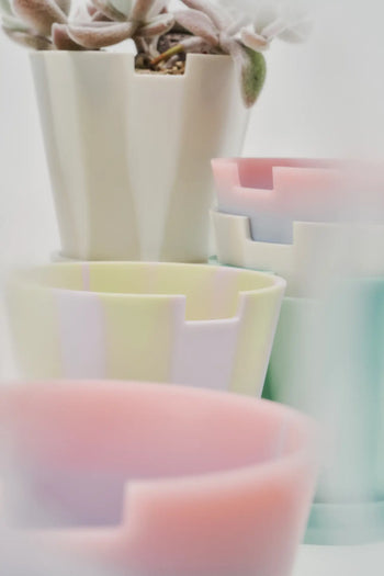 A soft-focus image displays several Plastic Ark Dirty Pots by Plastic Ark in pastel colors, some stacked with geometric edges. A plant with fuzzy leaves sits in the top pot, creating a light and airy vibe.