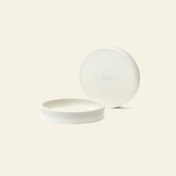 The Plastic Ark Dirty Pot is a simple, round, white recycled plastic pot. Its matching lid, embossed with Plastic Ark, rests slightly ajar behind it on a plain, light background.