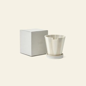 The Plastic Ark Dirty Pot by Plastic Ark, a white, modern, angular cup with matching round saucer, is shown beside a plain light grey box on a cream background—resembling a unique plant pot made from recycled plastic.