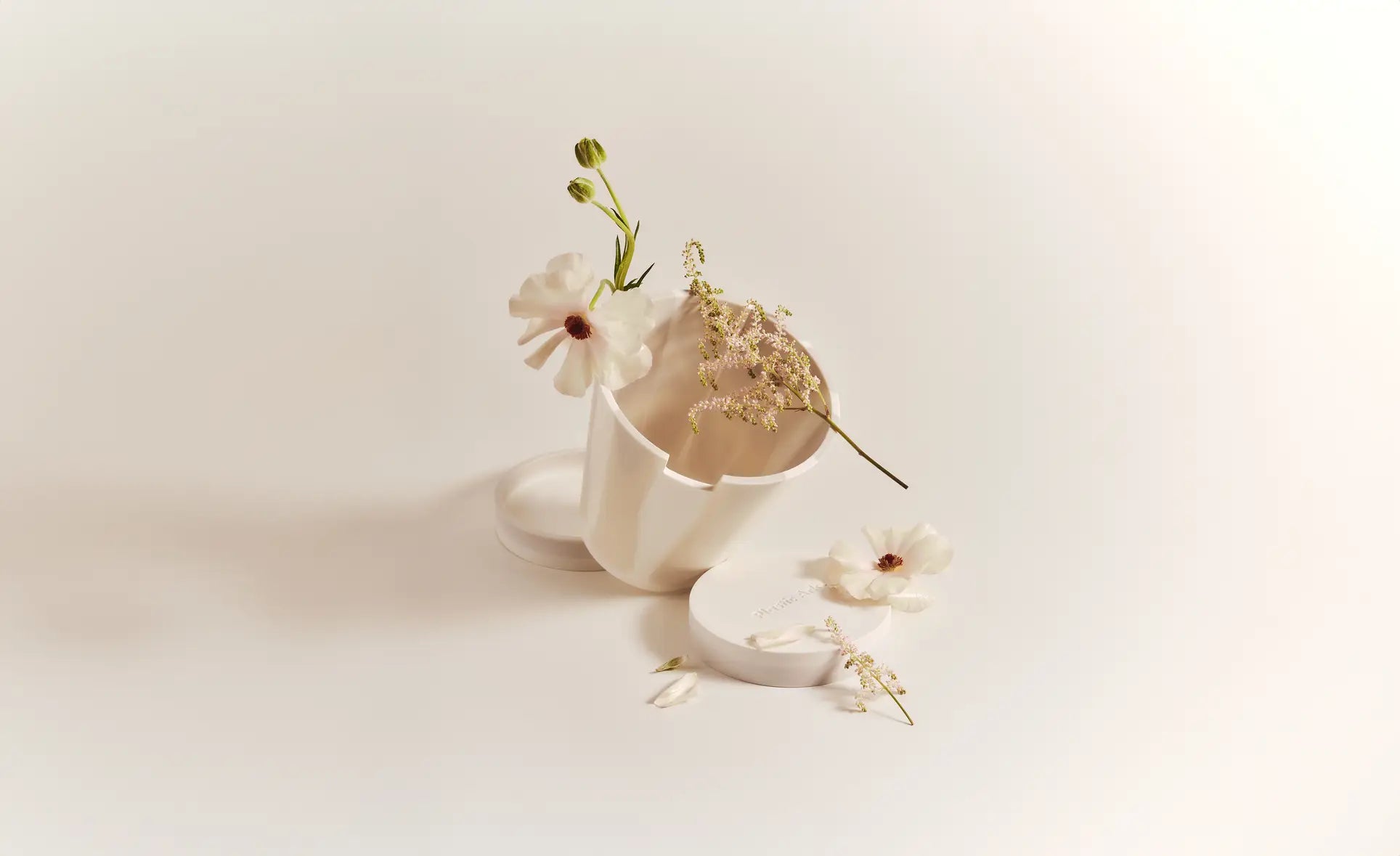 A Plastic Ark Dirty Pot by Plastic Ark lies on its side with its lid off, surrounded by delicate white flowers and sprigs on a minimal light background—perfect inspiration for displaying this unique sustainable plant pot.