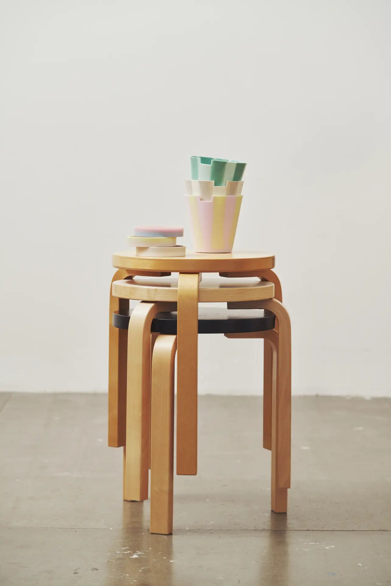 Three wooden stools are stacked on a gray concrete floor against a plain white wall, topped with pastel cups, plates, and the Plastic Ark Dirty Pot by Plastic Ark.