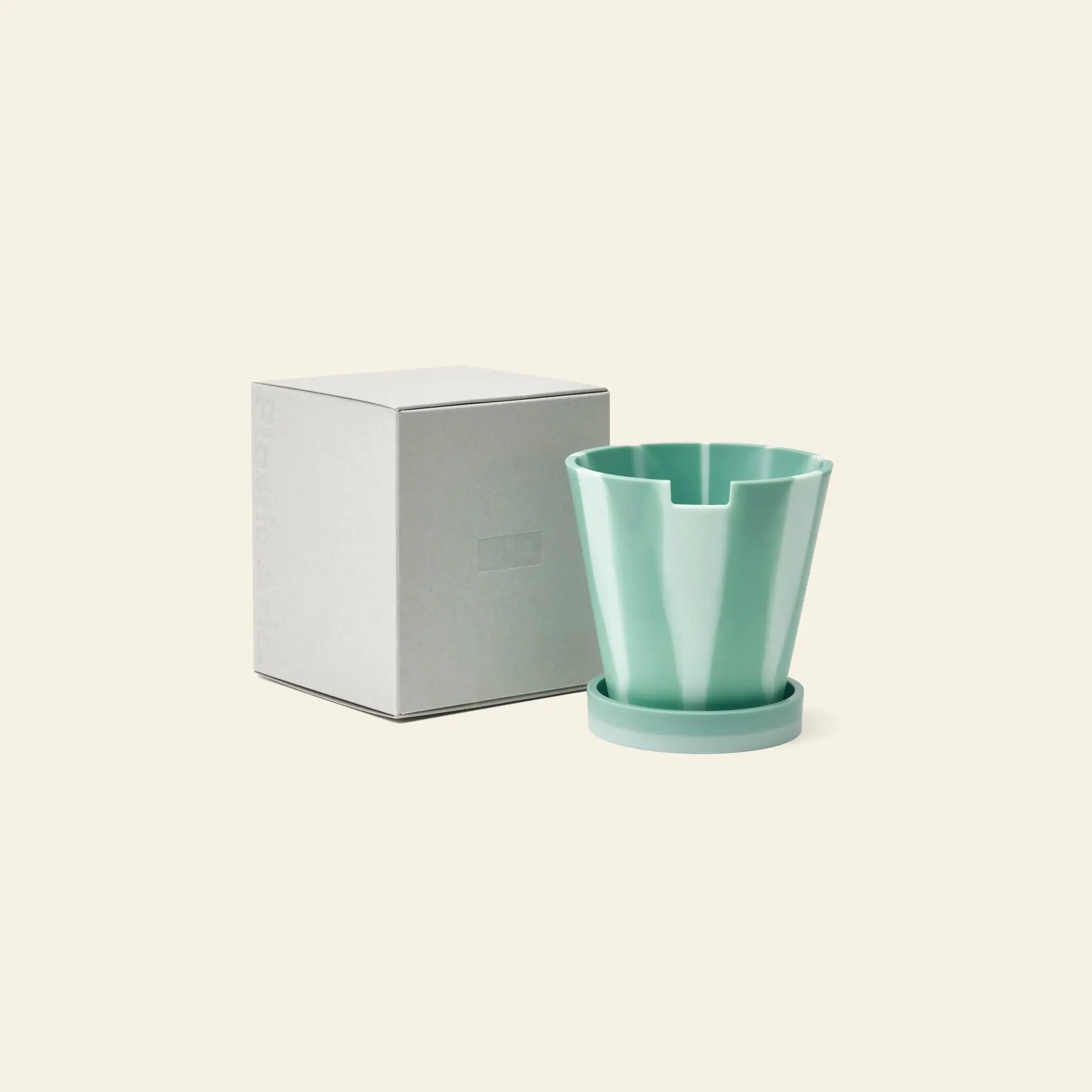 The Plastic Ark Dirty Pot by Plastic Ark, a light green geometric plant pot with matching saucer, is shown beside a plain square white box on a beige background.