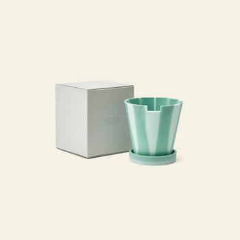 The Plastic Ark Dirty Pot by Plastic Ark, a light green geometric plant pot with matching saucer, is shown beside a plain square white box on a beige background.