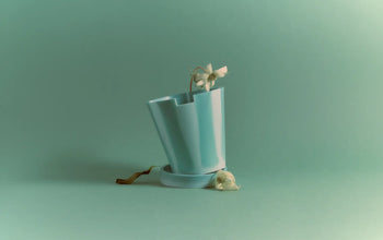 The Plastic Ark Dirty Pot by Plastic Ark is a turquoise, tilted sustainable plant pot holding a wilted white flower, with another flower beside it, all set against a matching turquoise background.