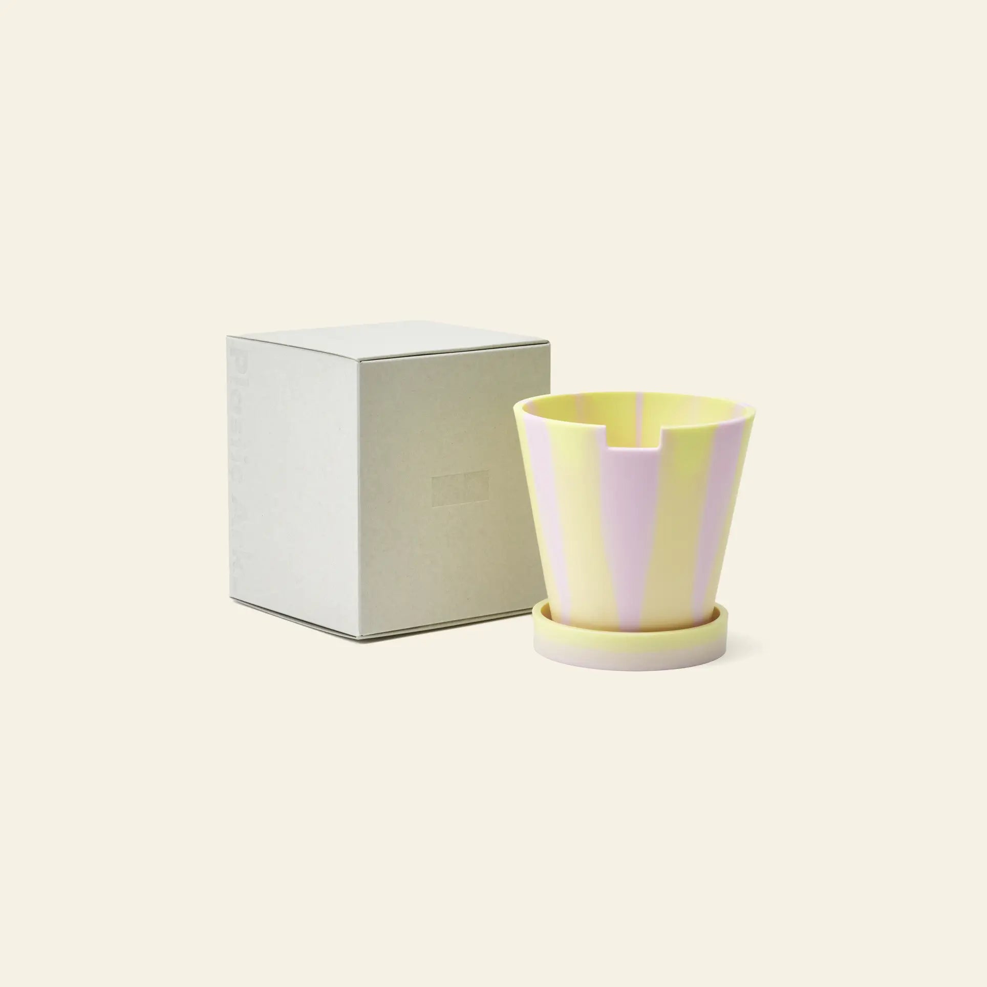 The Plastic Ark Dirty Pot by Plastic Ark, featuring pastel yellow and lavender stripes and a matching saucer made from recycled plastic, is placed beside a plain light gray box on a cream-colored background.