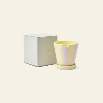 The Plastic Ark Dirty Pot by Plastic Ark, featuring pastel yellow and lavender stripes and a matching saucer made from recycled plastic, is placed beside a plain light gray box on a cream-colored background.