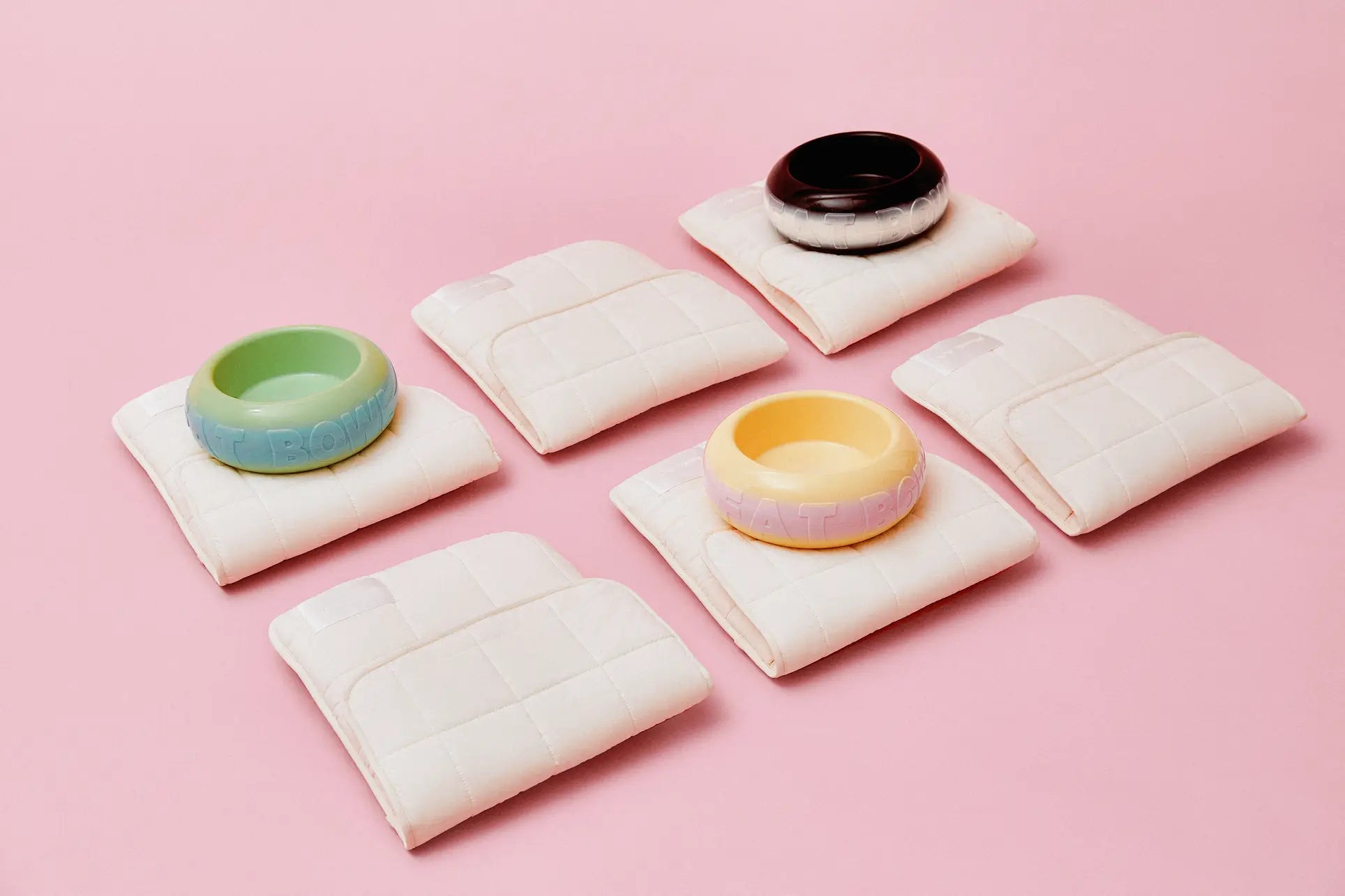 Six folded white quilted mats are displayed on pink, each holding a Plastic Ark Fat Bowl by Plastic Ark—colorful, round, and made from recycled plastic in green, blue, black, yellow, or pastel for a customizable touch in any space.