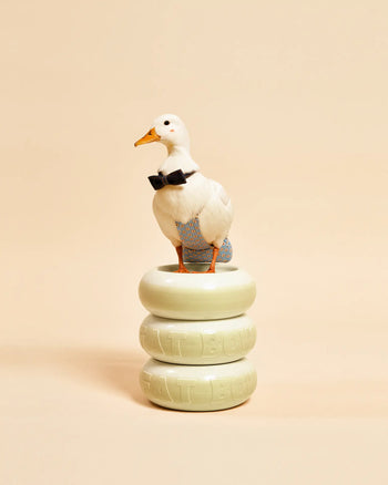 A white duck in a black bow tie and blue pants stands on two stacked Plastic Ark Fat Bowls by Plastic Ark, embossed with FAT BOWL and featuring an elegant modular design, set against a plain beige background.