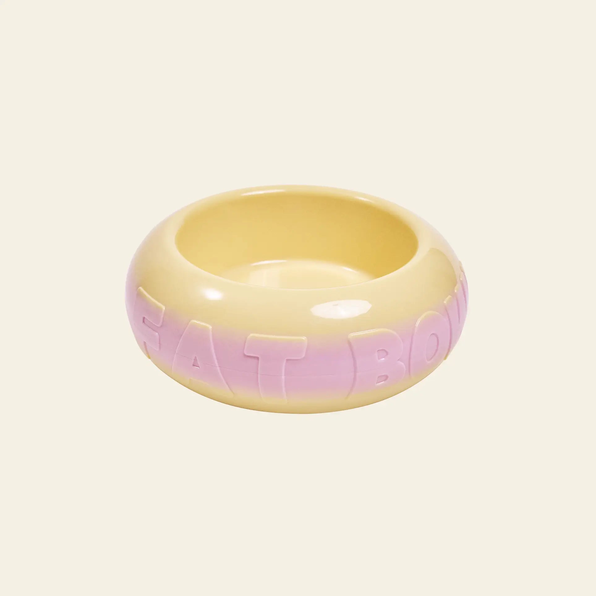 The Plastic Ark Fat Bowl by Plastic Ark is a round, glossy ceramic pet bowl featuring a yellow and light pink gradient and large raised “FAT BOWL” lettering. Inspired by modular design, it’s shown against a plain beige background.
