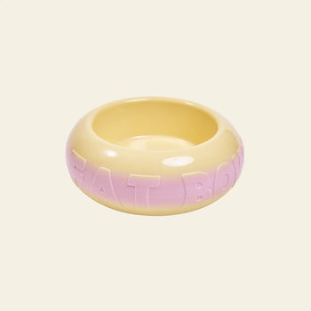 The Plastic Ark Fat Bowl by Plastic Ark is a round, glossy ceramic pet bowl featuring a yellow and light pink gradient and large raised “FAT BOWL” lettering. Inspired by modular design, it’s shown against a plain beige background.