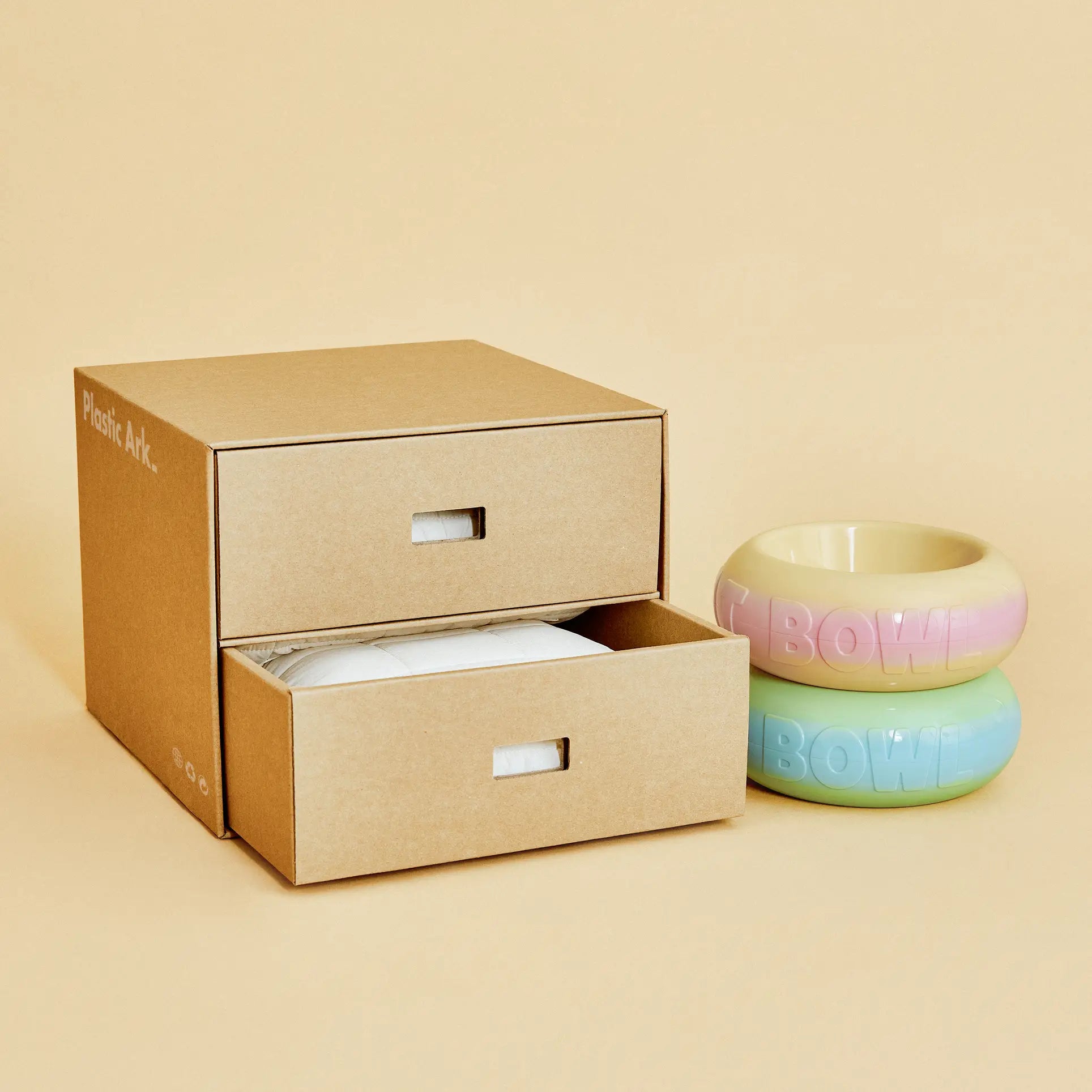 A modular cardboard storage box with two open drawers sits next to Plastic Ark Fat Bowls by Plastic Ark—one in pink and yellow, the other in blue and green—displayed on a beige background.