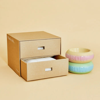 A modular cardboard storage box with two open drawers sits next to Plastic Ark Fat Bowls by Plastic Ark—one in pink and yellow, the other in blue and green—displayed on a beige background.