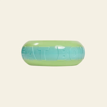 The Plastic Ark Fat Bowl by Plastic Ark is a round, gradient pet bowl in green-to-blue plastic with embossed FAT BOWL lettering, inspired by modular design and photographed against a plain beige background.