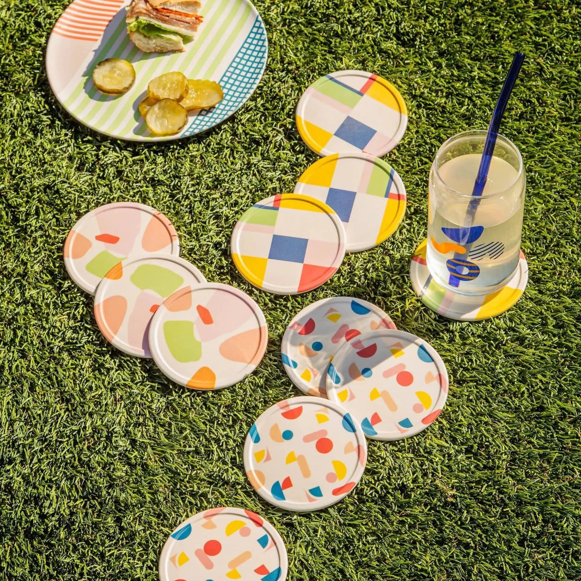 Poketo Bamboo Coasters, Set of 4, add a playful touch to your picnic setup—colorful patterns arranged on grass with a drink and sandwich in the background for a bright, inviting tabletop scene.