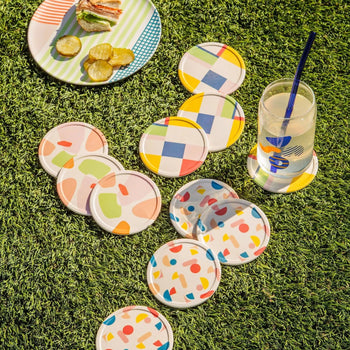Poketo Bamboo Coasters, Set of 4, add a playful touch to your picnic setup—colorful patterns arranged on grass with a drink and sandwich in the background for a bright, inviting tabletop scene.