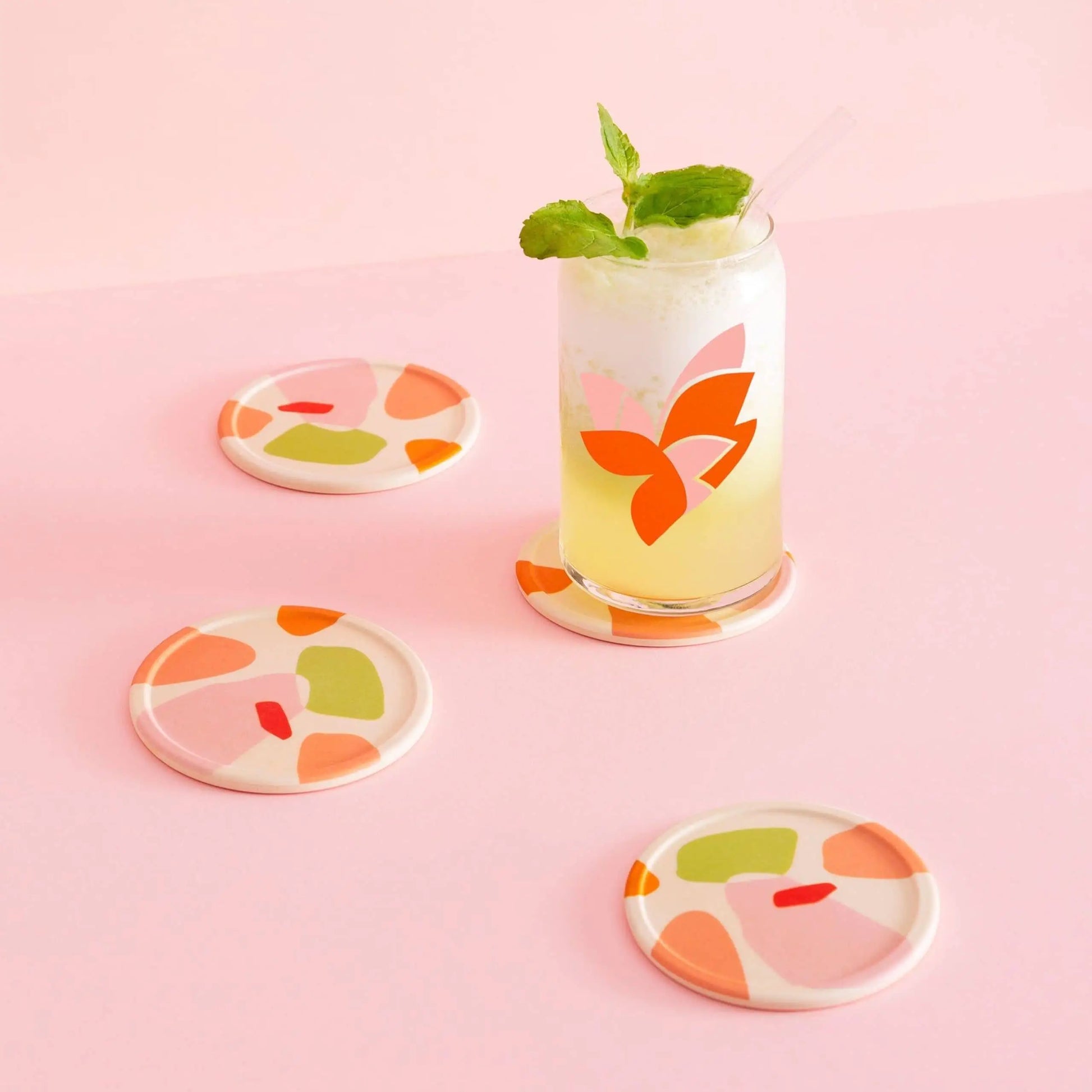 A glass of yellow lemonade with ice, mint, and a straw rests on a Poketo Bamboo Coaster. Three more coasters from the set of four feature abstract designs that add color to the cheerful pink tabletop.