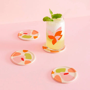 A glass of yellow lemonade with ice, mint, and a straw rests on a Poketo Bamboo Coaster. Three more coasters from the set of four feature abstract designs that add color to the cheerful pink tabletop.