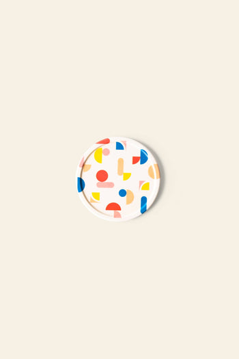 The Poketo Bamboo Coasters, Set of 4, feature colorful geometric shapes in red, yellow, blue, and pink on a round white surface, adding a vibrant touch to any tabletop display.