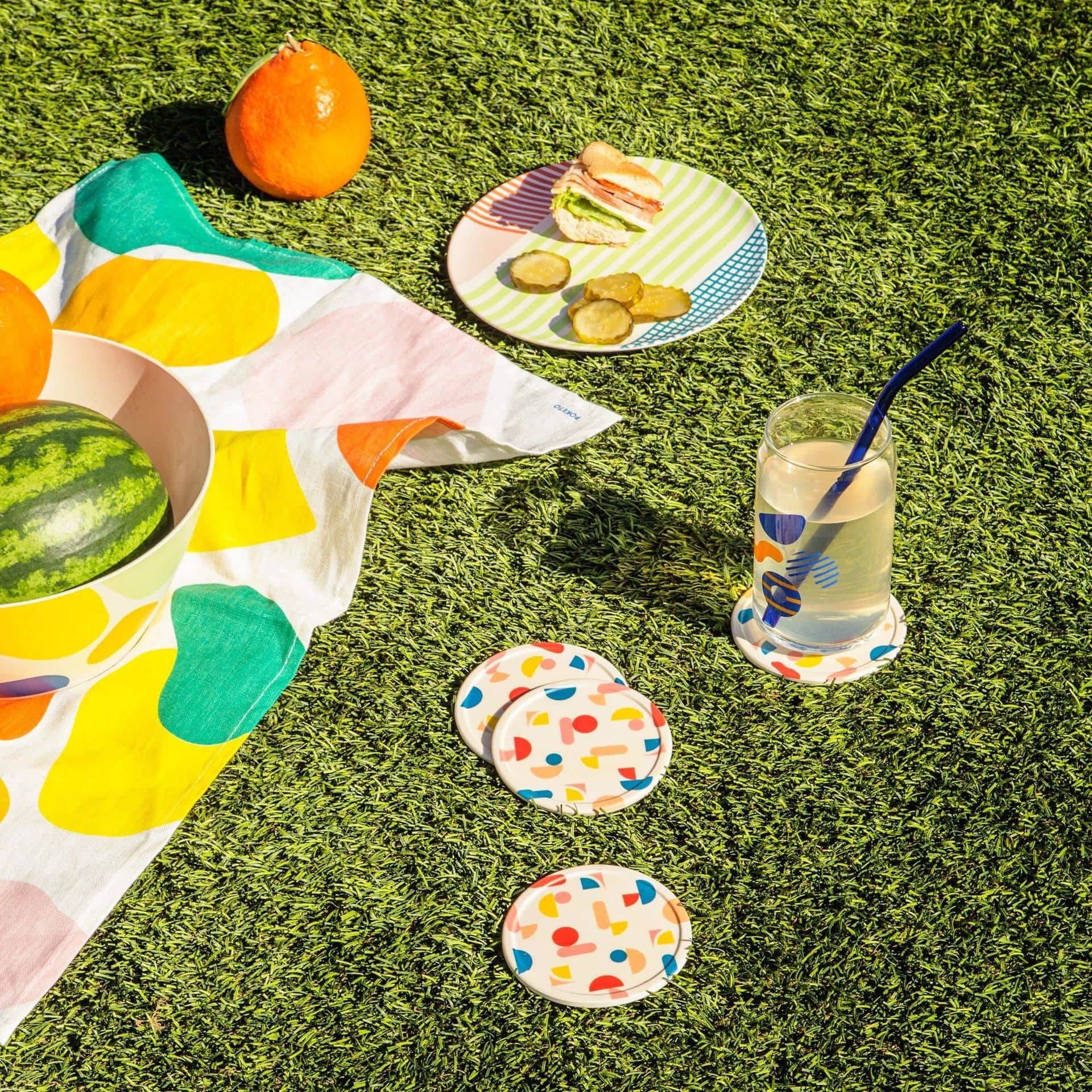 A vibrant picnic on green grass features Poketos Bamboo Coasters (Set of 4), a patterned cloth, fruit bowl, sandwiches with pickles, and lemonade—showcasing the coasters’ natural beauty in the sunlight.