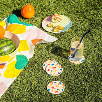 A vibrant picnic on green grass features Poketos Bamboo Coasters (Set of 4), a patterned cloth, fruit bowl, sandwiches with pickles, and lemonade—showcasing the coasters’ natural beauty in the sunlight.