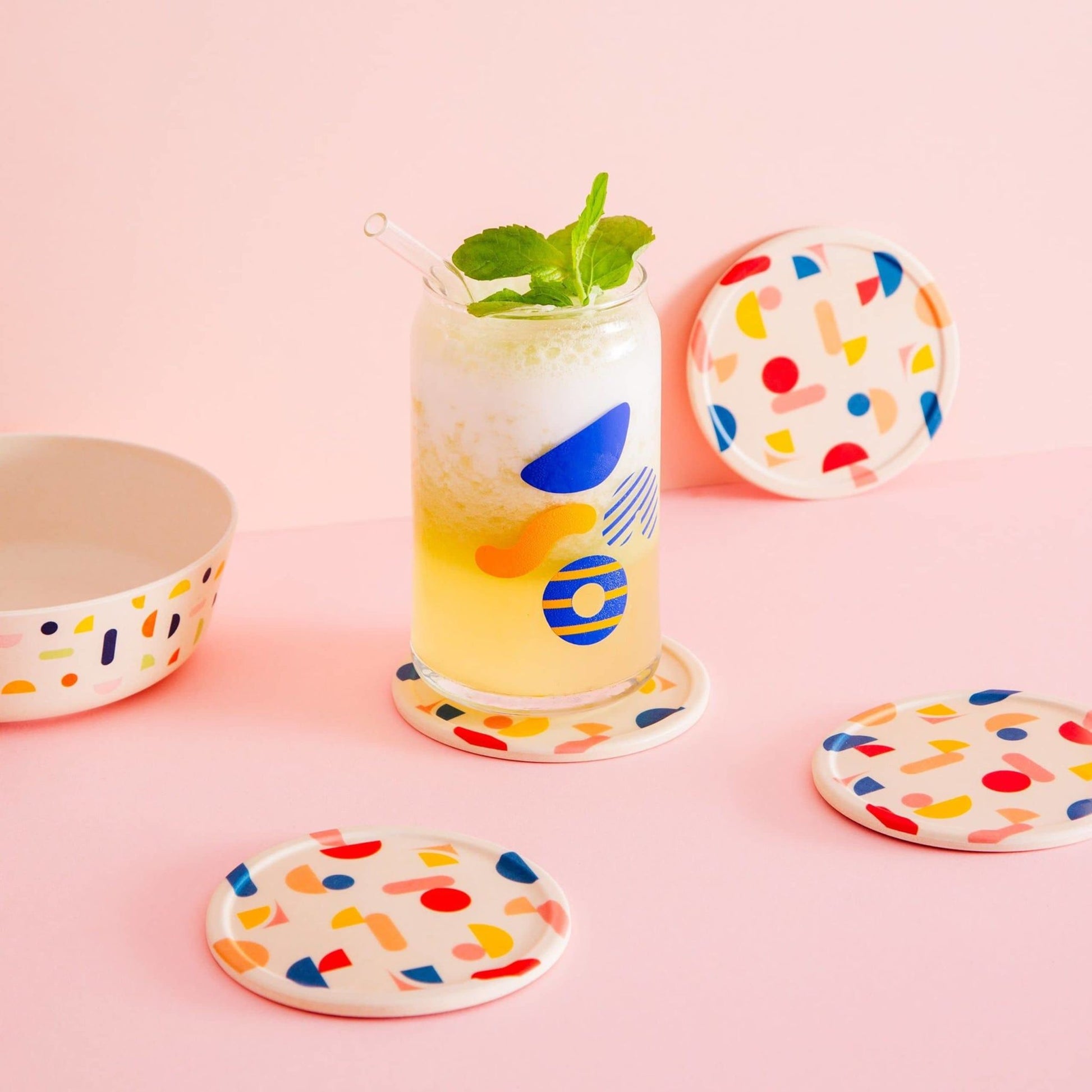 A glass of yellow and white frothy drink with a straw and mint sits on a Poketo Bamboo Coasters, Set of 4. Matching coasters and bowl accent the pink tabletop, all set against a pink background.