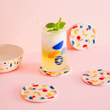 A glass of yellow and white frothy drink with a straw and mint sits on a Poketo Bamboo Coasters, Set of 4. Matching coasters and bowl accent the pink tabletop, all set against a pink background.