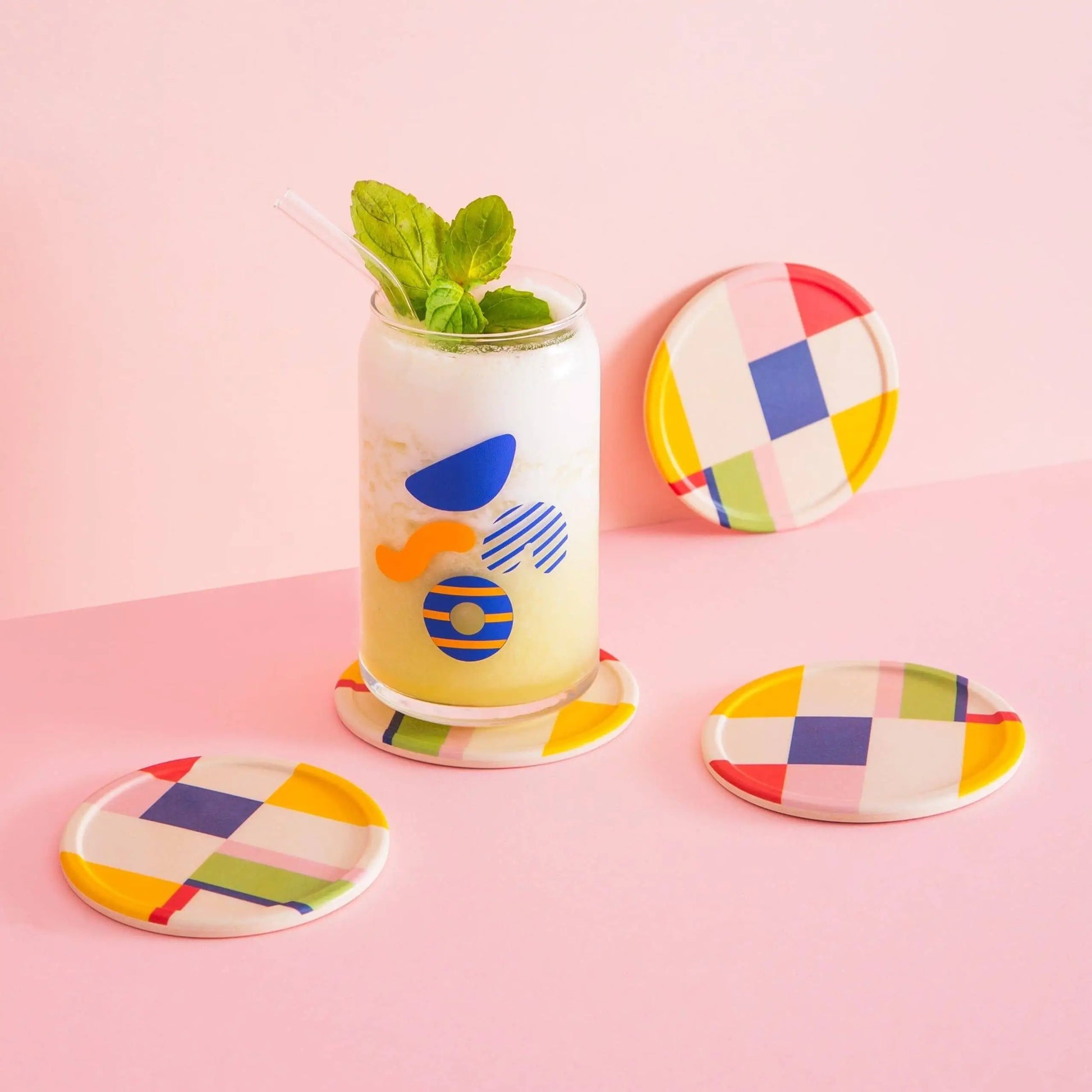 A glass of a light yellow drink sits on a Poketo Bamboo Coaster from the Set of 4, featuring colorful geometric patterns. Three matching coasters are nearby, all on a pink tabletop.