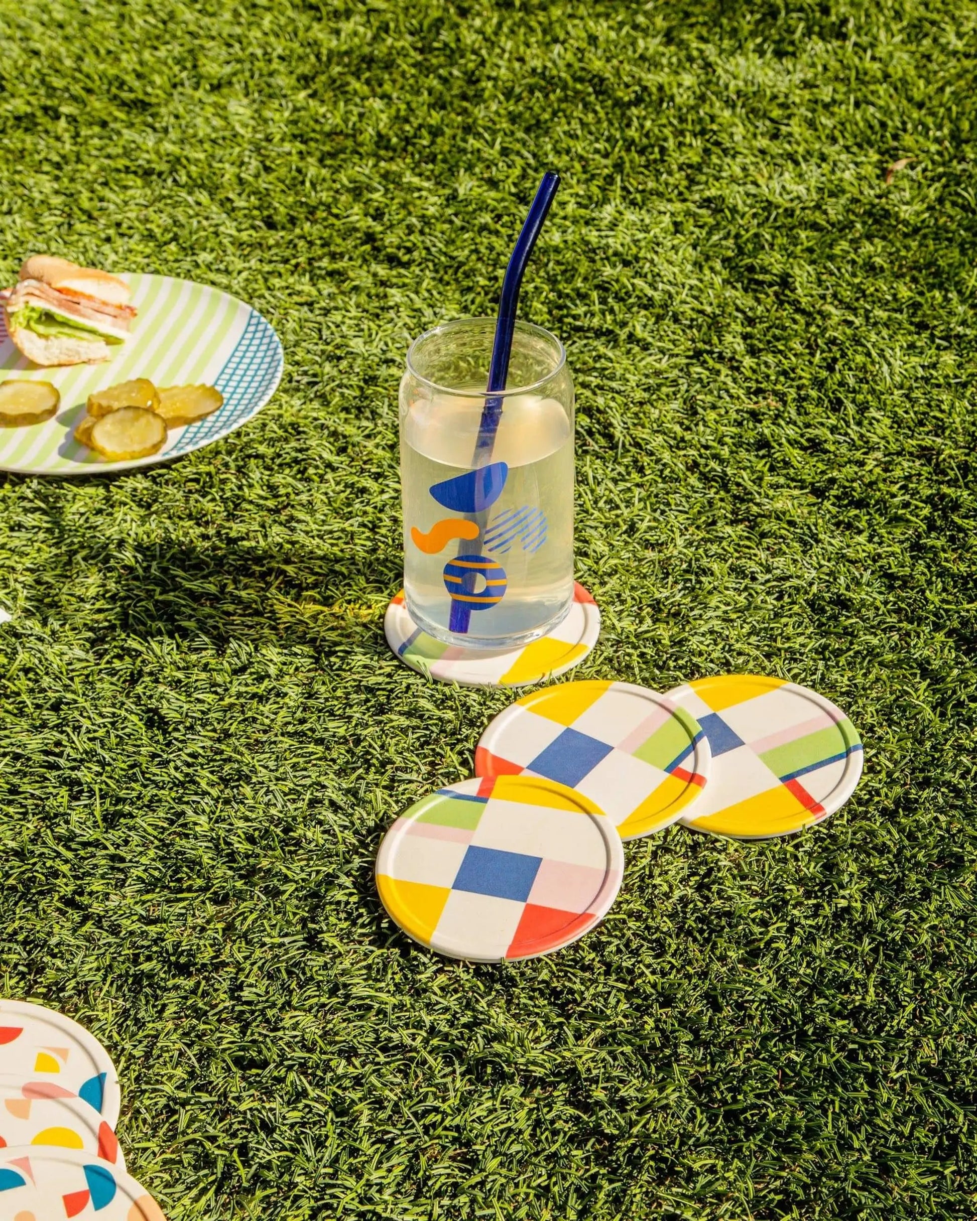 A glass of lemonade with a blue straw sits on Poketos Bamboo Coasters, Set of 4, arranged on green grass. Nearby, a plate with crackers and pickles suggests an inviting picnic atmosphere.