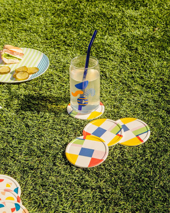 A glass of lemonade with a blue straw sits on Poketos Bamboo Coasters, Set of 4, arranged on green grass. Nearby, a plate with crackers and pickles suggests an inviting picnic atmosphere.