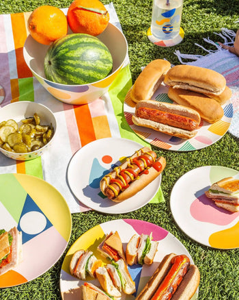 A vibrant picnic with hot dogs, sandwiches, watermelon, pickles, and drinks served on Poketo Bamboo Dinner Plates by Poketo atop a striped cloth on sunlit grass—ideal for eco-friendly gatherings.