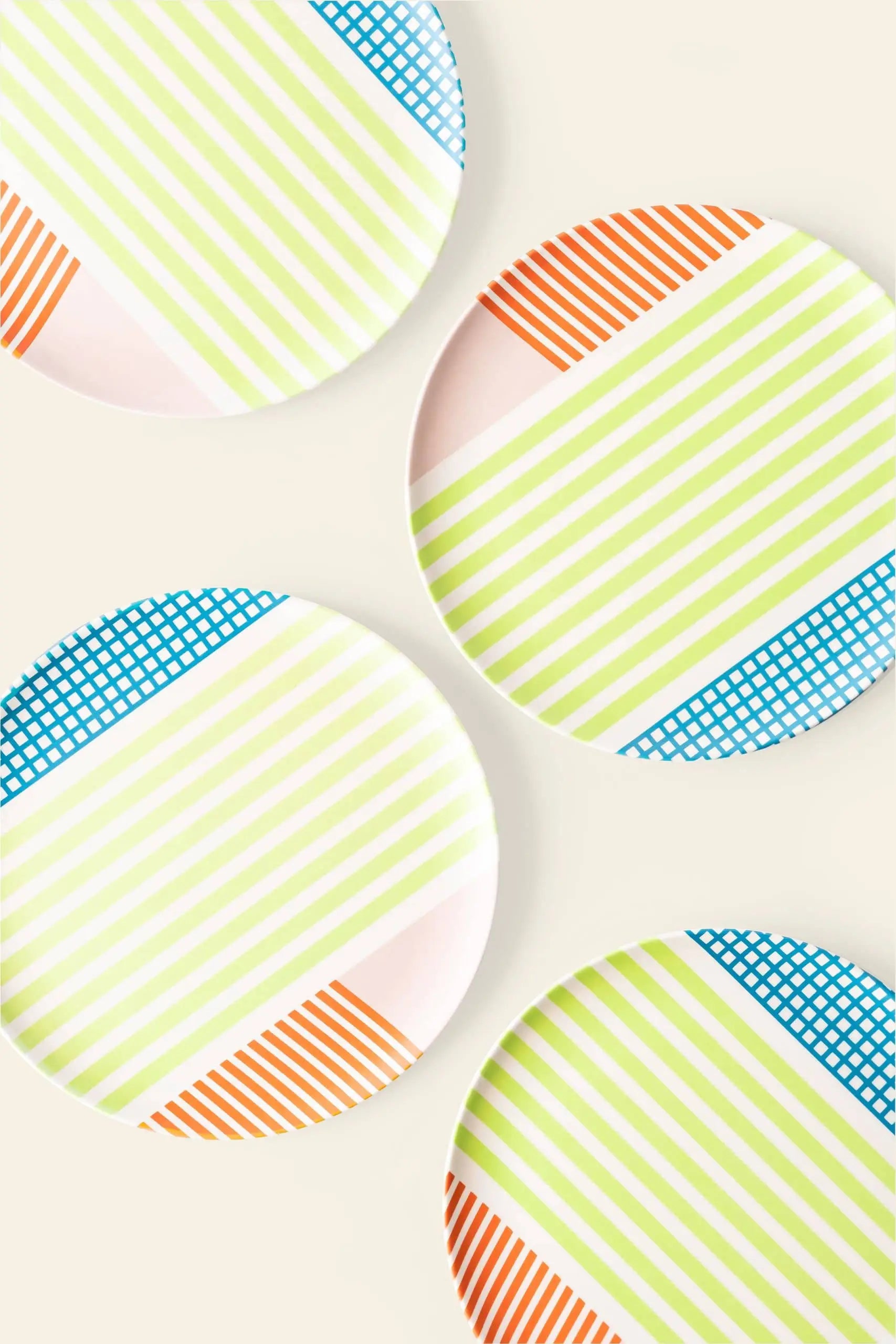 Four Poketo Bamboo Salad Plates by Poketo, featuring white surfaces with green stripes, orange lines, and blue grid patterns, are arranged on a light surface. The eco-friendly plates overlap slightly and sit at various angles.