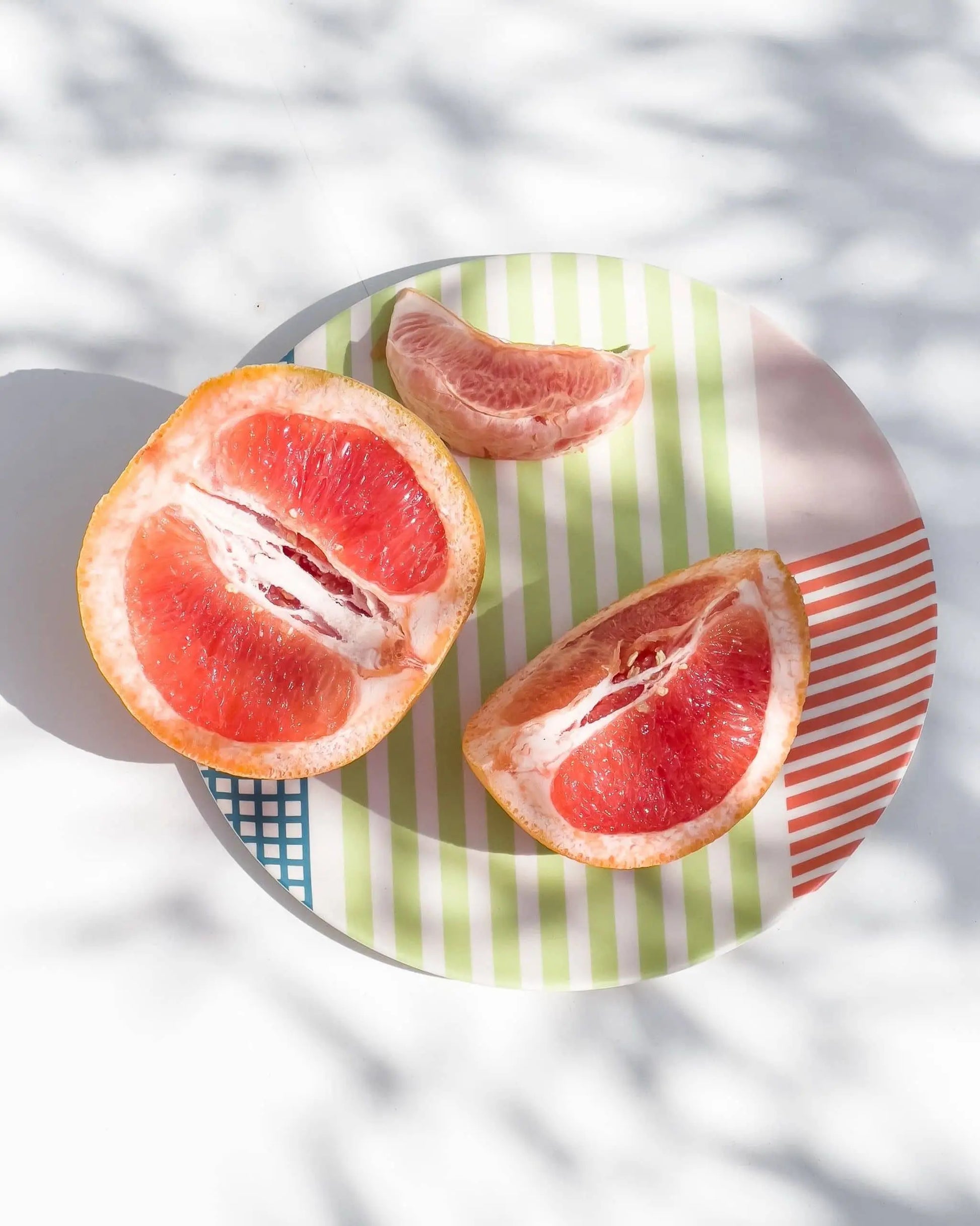 The Poketo Bamboo Salad Plate by Poketo holds two grapefruit halves and a wedge on a white surface in soft, dappled sunlight—an eco-friendly choice for sustainable dining.