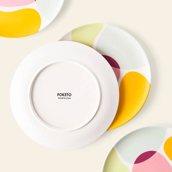 Three Poketo Bamboo Salad Plates display colorful abstract designs in yellow, pink, green, and burgundy on a cream surface. One plate is flipped to reveal the Poketo logo, emphasizing eco-friendly tableware for sustainable dining.