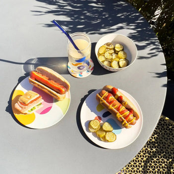 A round table set outdoors in bright sunlight features Poketo Bamboo Salad Plates by Poketo with hot dogs dressed with ketchup and mustard, a sandwich, pickles in a bowl, and lemonade served with a blue straw.