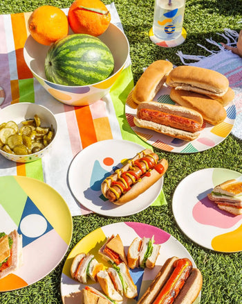A vibrant picnic on grass displays hot dogs, sandwiches, pickles, oranges, and watermelon served on Poketo Bamboo Salad Plates by Poketo—showcasing sustainable dining with eco-friendly tableware atop a striped cloth.