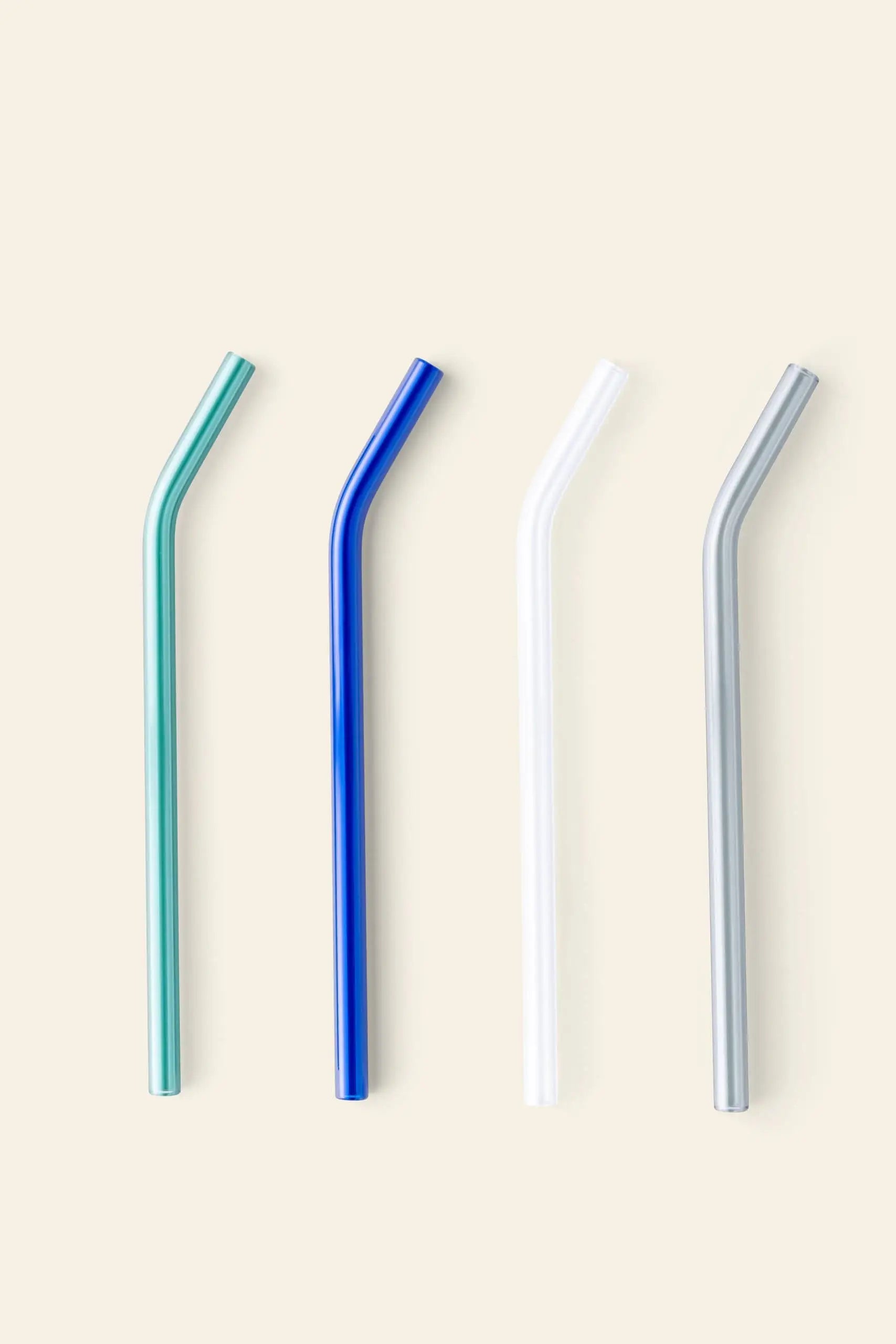 The Poketo Glass Straws, Set of 4 by Poketo features eco-friendly, reusable straws in teal, blue, white, and silver glass. Each straw is angled and displayed vertically on a simple light background.