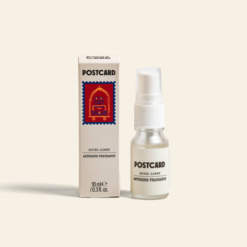 A 10ml bottle of Postcard Anywhere Fragrance – Hotel Lobby by Postcard is placed beside its box, which features a red postage stamp-inspired illustration, all set against a plain light background.