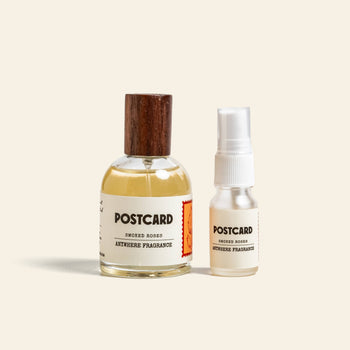 Two bottles of Postcard Anywhere Fragrance, 10ml - Smoked Roses by Postcard, with warm amber and rose notes, sit side by side—a larger bottle with a wooden cap and a smaller spray—on a light beige background.