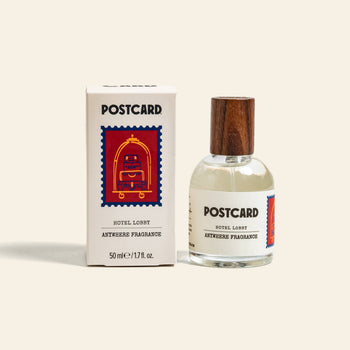 A 50ml bottle of Postcard Anywhere Fragrance – Hotel Lobby by Postcard sits beside its box, which displays an illustrated red luggage cart on a blue background.
