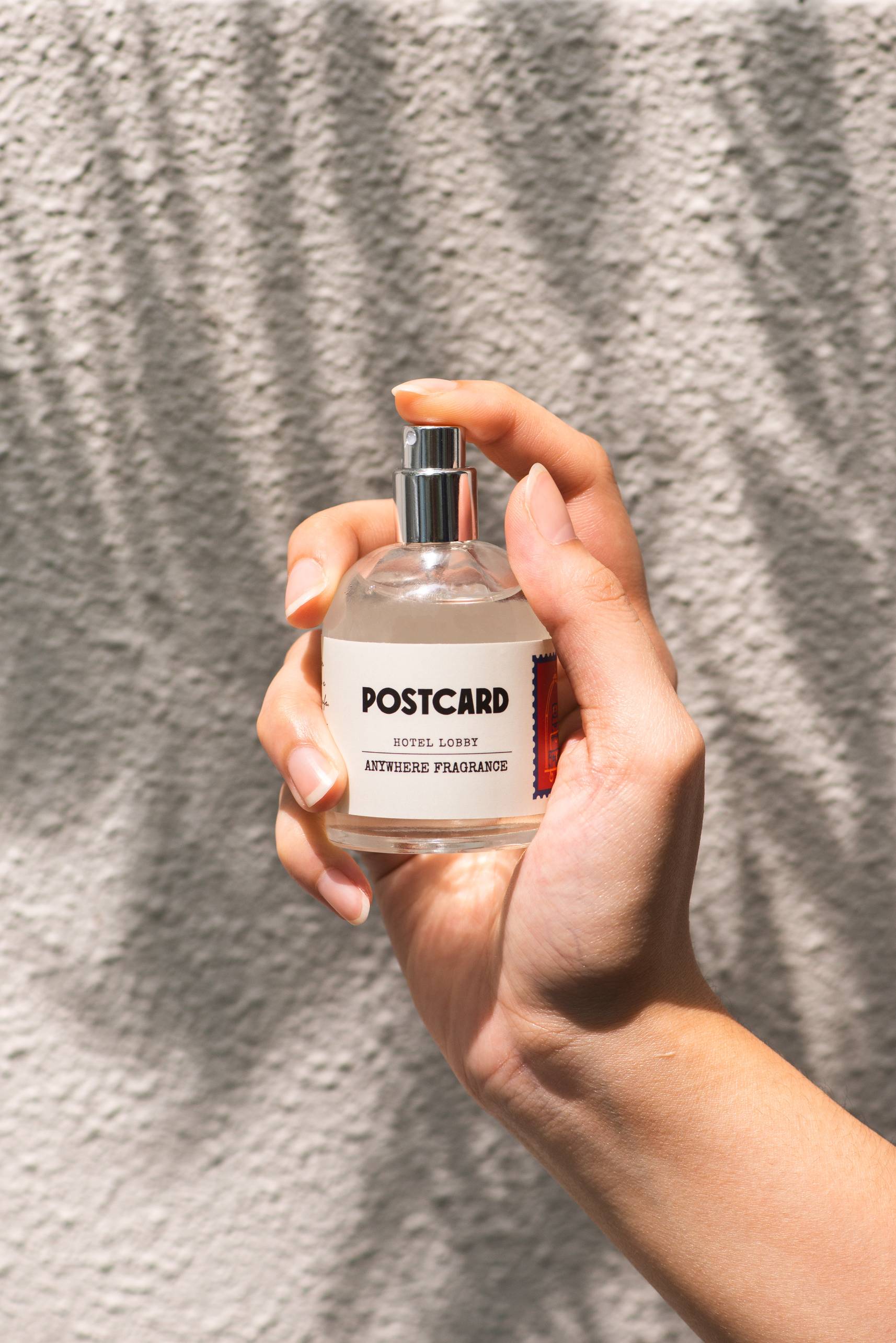 A hand holds a clear 50ml bottle of Postcard Anywhere Fragrance in the Hotel Lobby scent by Postcard, set against a sunlit, textured wall with soft shadows.