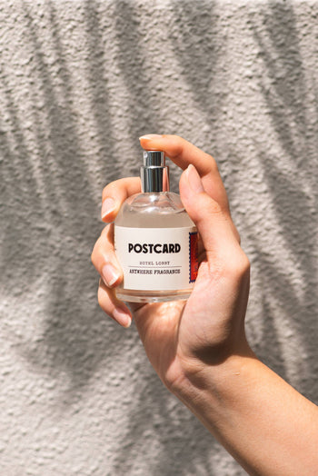 A hand holds a clear 50ml bottle of Postcard Anywhere Fragrance in the Hotel Lobby scent by Postcard, set against a sunlit, textured wall with soft shadows.