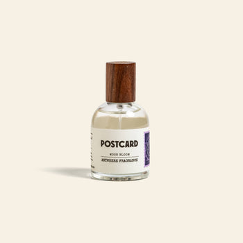 A clear 50ml Postcard Anywhere Fragrance—Moon Bloom—with a wooden cap, white label, and purple stamp graphic sits against a beige background, capturing dreamy scent notes in elegant glass.
