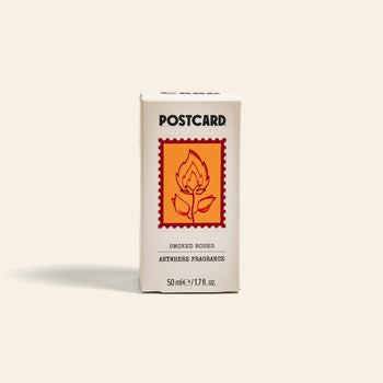 A beige box labeled Postcard Anywhere Fragrance, 50ml - Smoked Roses by Postcard features an orange stamp with a flaming rose and delivers amber fragrance with smoky rose perfume notes.