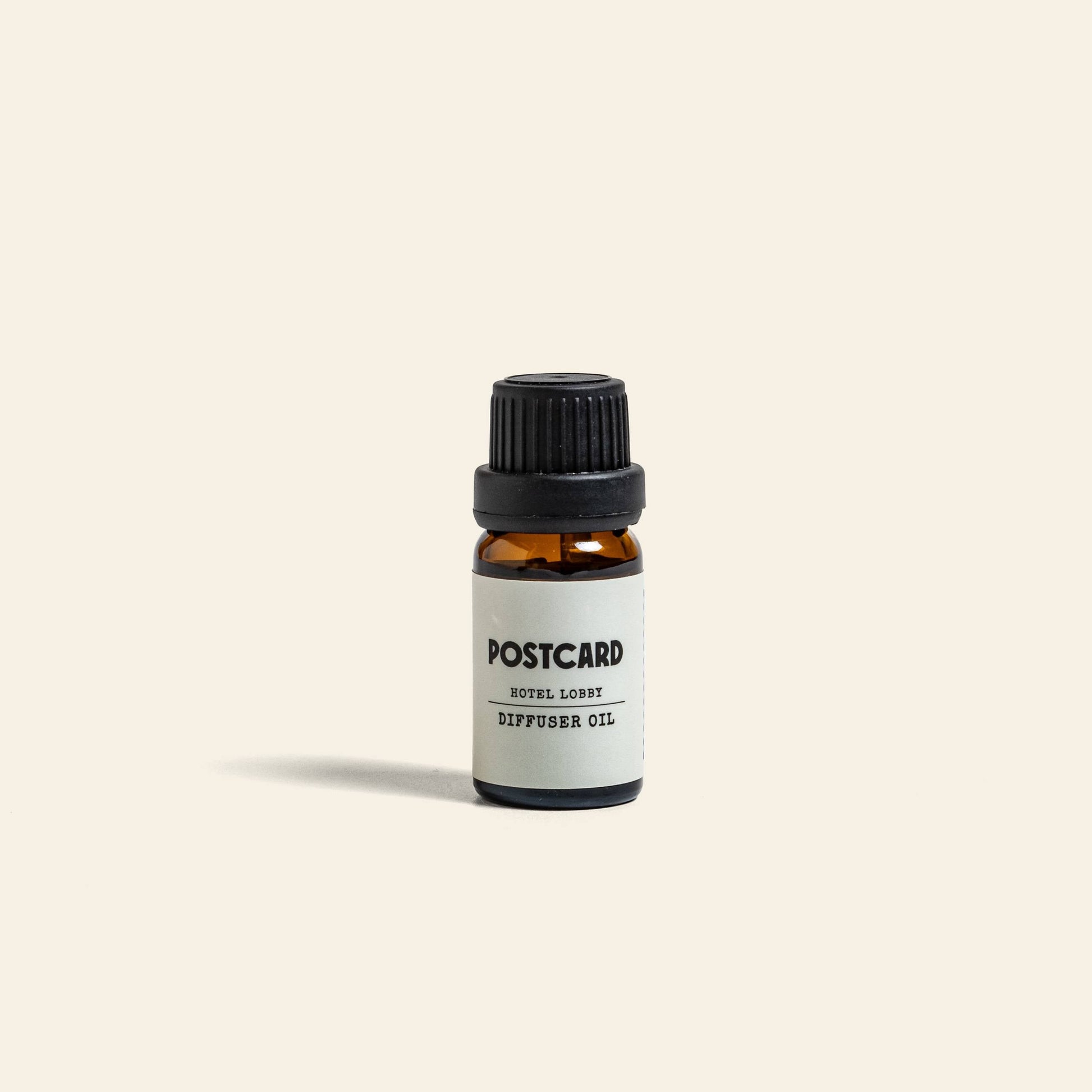 A small amber glass bottle with a black cap and a white Postcard label sits centered on a light beige background, showcasing Postcard Diffuser Oil, 10g - Hotel Lobby for an elegant, hotel-inspired experience.