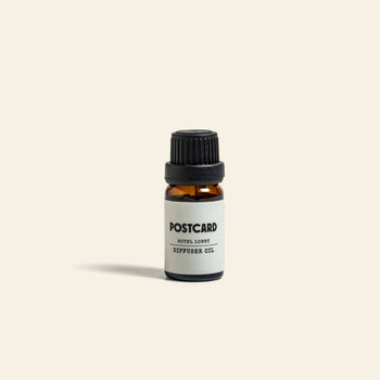 A small amber glass bottle with a black cap and a white Postcard label sits centered on a light beige background, showcasing Postcard Diffuser Oil, 10g - Hotel Lobby for an elegant, hotel-inspired experience.