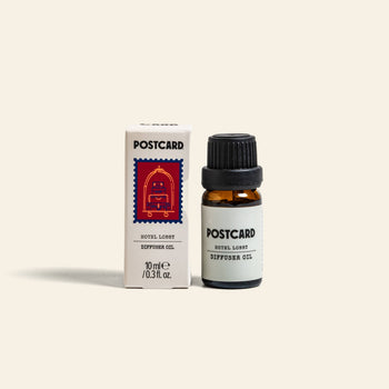 A 10g bottle of Postcard Diffuser Oil by Postcard stands beside its white box, which features a red postage stamp illustration. This luxe blend captures the essence of Hotel Lobby luxury in every drop.