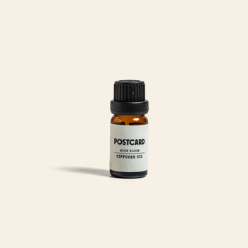 A small amber glass bottle with a black cap labeled “Postcard Diffuser Oil, 10g - Moon Bloom” by Postcard stands upright on a light beige background, offering calming aromas to enhance any space.
