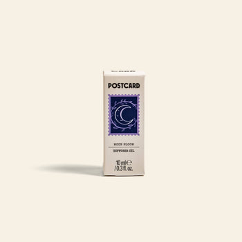 A small white Postcard box labeled Postcard Diffuser Oil, 10g - Moon Bloom, features a blue crescent moon and stars stamp. Calm aromas for your lunar lounge; the box stands upright on a light surface.