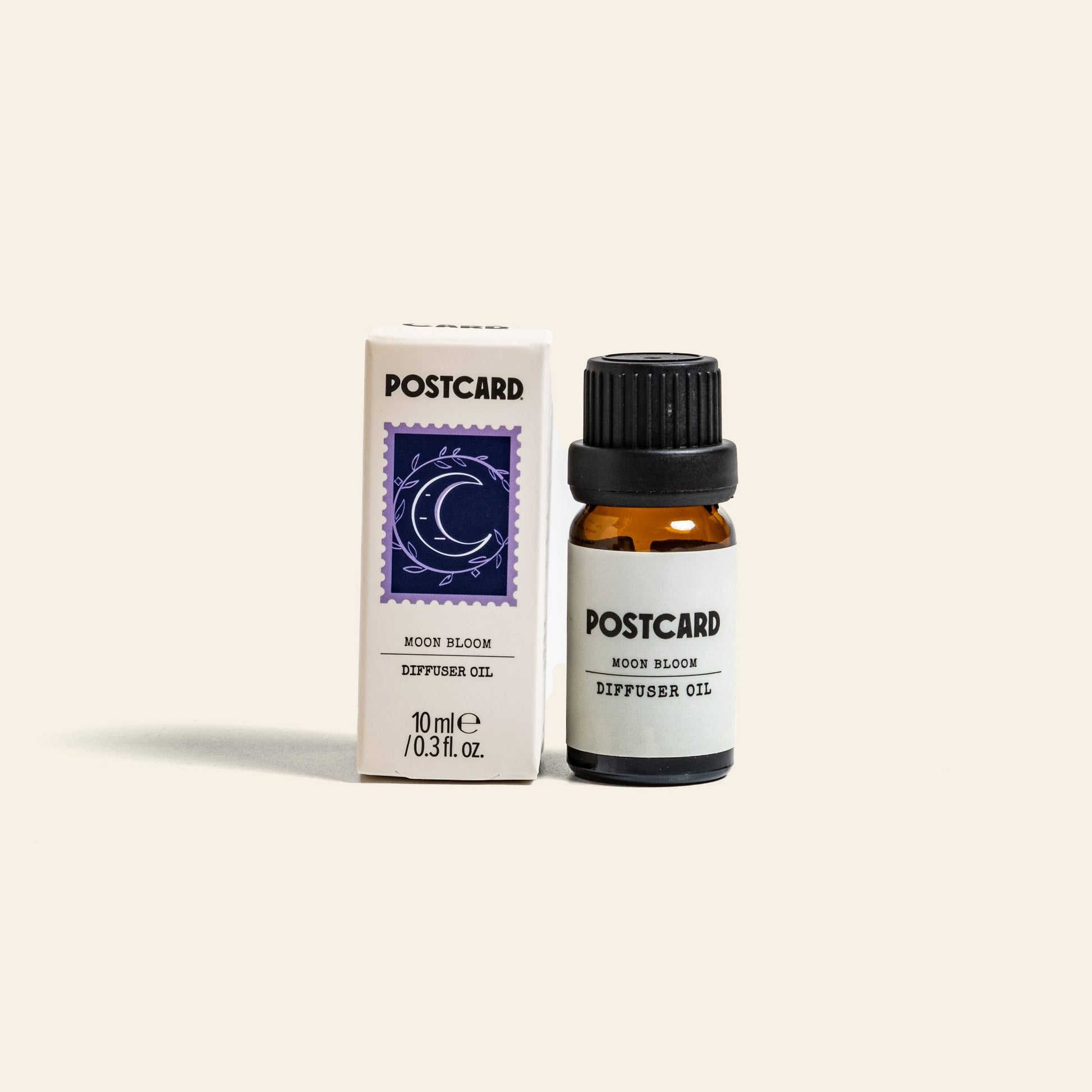 The Postcard Diffuser Oil, 10g - Moon Bloom by Postcard is shown in a small amber bottle next to its box, adorned with a blue stamp illustration of a crescent moon and stars, all set against a simple light background.
